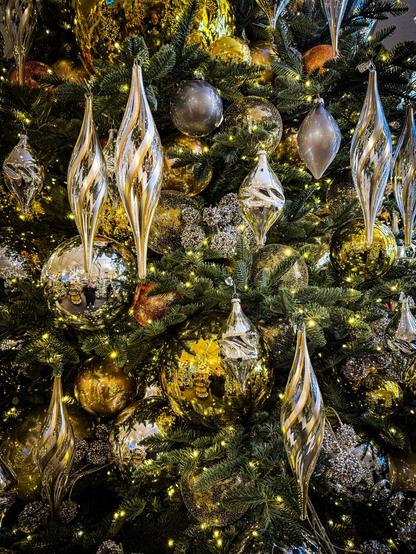 This is a close-up shot of a heavily decorated Christmas tree. The tree branches are a dark green and are adorned with a large number of ornaments in shades of gold and silver. Many of the ornaments are teardrop-shaped, appearing reflective and metallic, while others are spherical and textured. Small, warm-toned lights are scattered throughout the branches, creating a sparkling effect. The ornaments are densely packed, giving the tree a luxurious and festive appearance. Provided by @altbot, generated privately and locally using Gemma3:27b