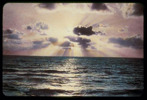 The image depicts a captivating sunset over the ocean, with rays of sunlight piercing through dark clouds. The sky is a mix of warm pink hues and cooler blue tones, creating a striking contrast that highlights the dramatic atmosphere of the scene. The waves on the water's surface reflect these colors, adding to the overall beauty of the composition. This picturesque moment seems to be captured in a personal photograph album or archive collection, as suggested by its size (2 x 2 inches) and the presence of an identification label from Matson Photo Service.