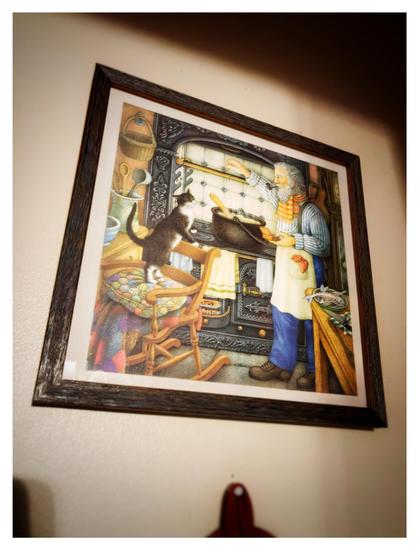 Photo, taken at an angle, of a wooden picture frame displaying a colorful illustration of a kitchen scene: an old bearded man in a scarf and apron sprinkling ingredients into a pot of bubbling stew, while a black-and-white tuxedo cat props herself up on the back of a nearby rocking chair and sniffs at the pot appreciatively. The picture hangs on a plain off-white wall with some shadows in a corner. Below the picture frame we can partially see an oven mitt hanging from a thumbtack.