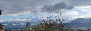 The photo is taken from some way up a steep path. Some nearby shrubs and the top of a street lamp are at the bottom of the picture. There are a lot of light grey but thick clouds in the sky, but a pale blue band appears lower down just above a row of snowy rugged peaks and then more light clouds appear between the mountains, at the level of some nearer dark hills. A black and white skyscraper breaks the view of the lower hills. There are town buildings at the bottom of the hills.