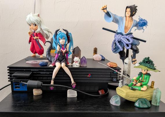 PlayStation 2 (Original Model) Laying horizontal ln a small black wooden table. Two figures on the top of the console, a QPosket Inuyasha figure and a sitting dragon Hatsune Miku figure. A Banpresto Sasuke Uchiha figure using his lightning blade from Shippuden on the right of the console, as well as a sitting Gon figure from Hunter x Hunter (on a rock, fishing, with a bird on his large round hat covered in moss). Various crystals surround the console and are on top of it.