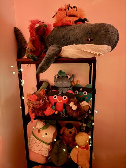 English: A shelf filled with a variety of colorful plushies, including a large blåhaj on top, and smaller creatures and characters below. The shelf is adorned with fairy lights, creating a cozy atmosphere. 

Deutsch: Ein Regal, das mit einer Vielzahl von bunten Plüschtieren gefüllt ist, darunter ein großer Blåhaj ganz oben und kleinere Plüschies darunter. Das Regal ist mit Lichterketten geschmückt und schafft eine gemütliche Atmosphäre.