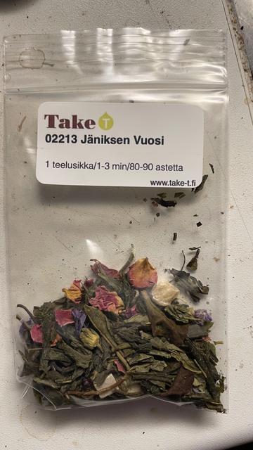 A small ziplock bag of tea leaves, flower petals and pieces of fruit and berries. The label on top states the name of the tea shop and the name of the tea blend. Below the names are brewing instructions in Finnish.