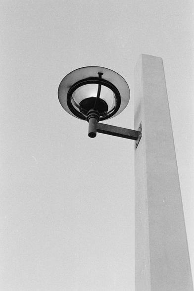 A black-and-white film photograph of a minimalist street lamp mounted on a tall rectangular concrete pillar. The composition emphasizes clean geometric shapes a circle, vertical rectangle, and straight lines against a pale, empty sky