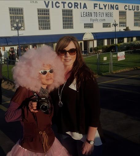 Two women posing for a photo in front of the Victoria Flying Club. I'm wearing a large pink afro wig, sunglasses, holding a camera, the Aimee is wearing sunglasses and a black cardigan.