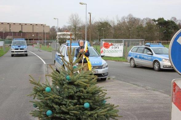 Im Hintergrund das Atommülllager Ahaus, auf der Zufahrtstraße 3 blau-weiße Polizeiautos. Hinten am Zaun ein Transparent gegen Atomkraft. In der Mitte vorne ein Weihnachtsbaum, geschmückt mit Brennelementekugel-großen-Christbaumkugeln, dahinter eine Demonstrant_in mit Atomkraft-Nein-Danke-Fahne