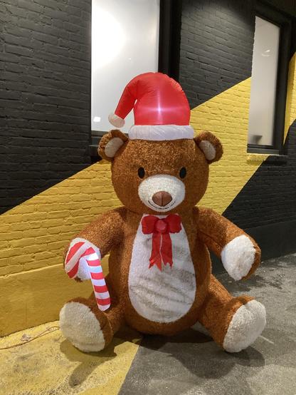 A large, inflatable teddy bear is wearing a red Santa hat, with a red bow tie and holding a striped candy cane. The bear is placed against a black and yellow wall.