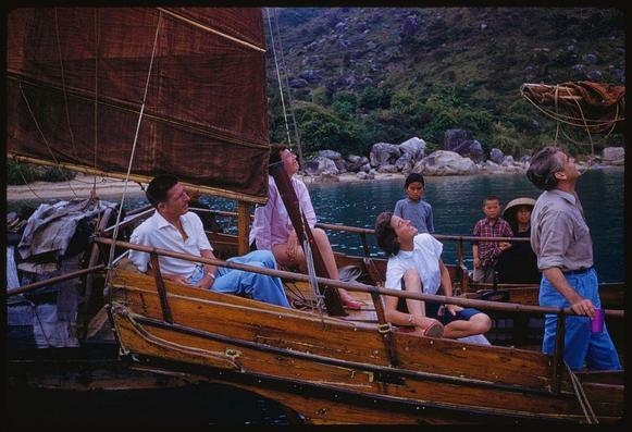 The image features a group of people on wooden boats in a body of water. The backdrop includes lush green hills, and the scene suggests an outdoor recreational activity or excursion in Hong Kong during 1959 as indicated by caption details.

Key elements include: multiple individuals sitting casually on boat benches; some adults are gazing upwards with expressions of wonder or enjoyment while others look around their surroundings attentively. There is a mixture of genders, ages and ethnicities among the group which comprises both men and women dressed in various styles reflecting mid-20th century fashion.

The setting appears to be during daylight hours under partly cloudy skies as inferred from lighting conditions on subjects' faces and objects within the scene like boats and water body. The color tone is primarily natural but shows signs of being a photograph taken with slide film which may affect its saturation levels in comparison to modern digital images or other formats.

The background's green hills suggest proximity to nature amidst urban development, hinting at Hong Kong's unique blend where city life coexists alongside lush landscapes. This juxtaposition might also be symbolic of the lifestyle and environment experienced during that era within this specific location known as "X5826," which presumably refers to a particular place or reference number in archives re [...]