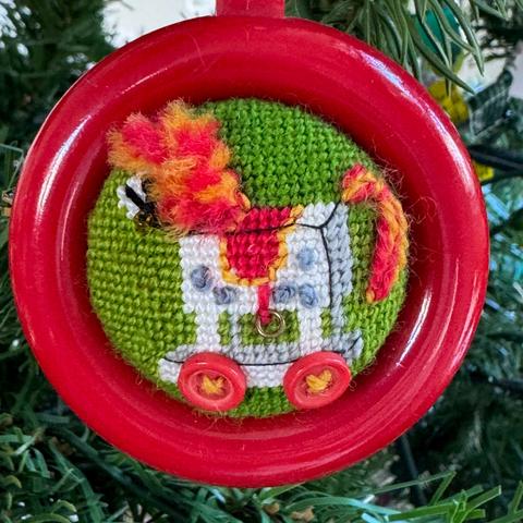 A round Christmas tree ornament. A small needlepoint picture of child's horse on red wheels (buttons). The horse is white with gray polkadots, his tale and mane are red and yellow. He also has a red and yellow saddle, all on a green background. The needlepoint is inside a red, round plastic frame, and the needlepoint is wrapped around half a sphere.