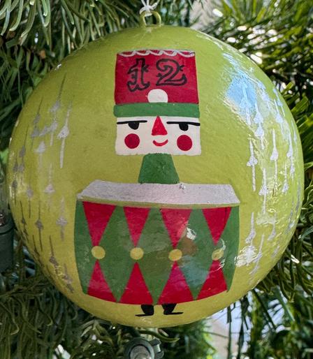 A round paper mache ornament with a light avocado green background and a drummer who has a square head and hat. The hat is red and green with the number "12" on it. There is a large drum in front of the drummer which is green with red triangles and gold dots. There are silver objects around the drummer which are straight lines with a spade in the center. The drummer has a red triangular nose, and round red cheeks.