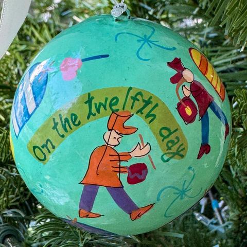 A round paper mache ornament with a turquoise background and various drummers. An avocado green banner that says, "On the twelfth day" is above one of the drummers who has an orange jacket and hat on with purple pants and orange shoes. He is carrying a small drum. To the side of him is another drummer dressed in maroon and dark blue, also with a drum.