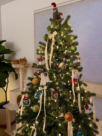 A view of the entire Christmas tree with all of the 12 Days of Christmas ornaments plus other handmade ornaments. On top of the tree is a sloth in a Santa hat, clinging to the top branch. White satin bows with long streamers decorate the tree. In the background is a cat tree with my kitten Olly on top. He has long orange hair, and he is looking at the tree. A large ficus tree is behind the cat tree. A large window with shades closed is behind the tree.