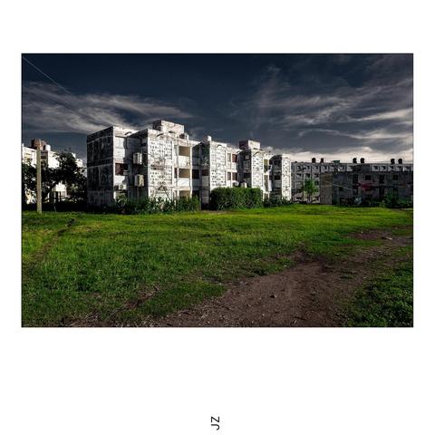 A concrete housing complex surrounded by green grass and illuminated by the sun. In the foreground a farm track and in the background a little pony. The sky is tinted in a faded dramatic dark blue tone.
