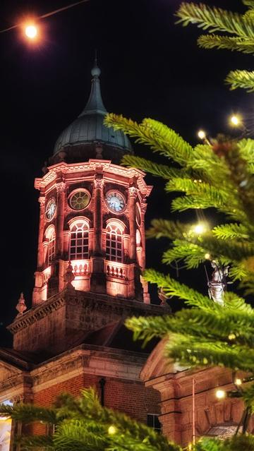 A vertical night photograph capturing the ornate clock tower of Dublin Castle, illuminated by warm, reddish-orange uplighting. The tower features a dome and circular clock faces visible through arched windows. The foreground is partially framed by out-of-focus green boughs of a Christmas tree adorned with warm white fairy lights, creating a strong festive atmosphere. A small statue is visible near the upper right section of the tower.