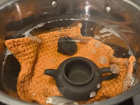 A dark Yixing teapot submerged in a pot of water on top of an orange tea towel.