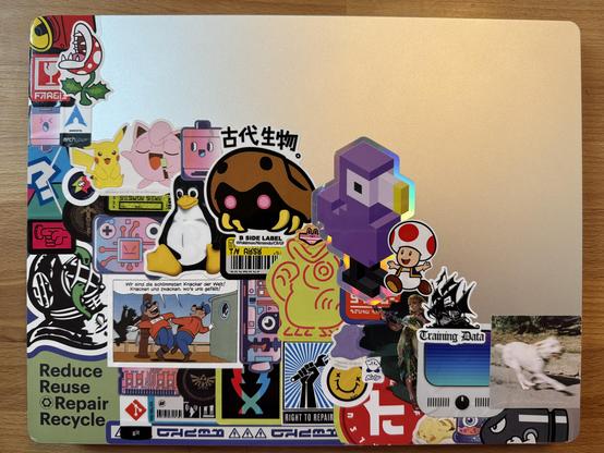 A picture showing my laptops lid, which is 2/3 covered with stickers, arranged in a way that they are spreading out from the bottom left corner. One of them is the „training data“ sticker mentioned in the post.