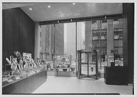 The image depicts a vintage, black and white photograph of an interior space resembling a department store or upscale retail establishment. The scene captures the view from behind a glass display case on 51st Street in New York City's Rockefeller Center district during June 9th, 1948.

Inside the building, there is a variety of merchandise displayed prominently within and through large glass windows. Items such as floral arrangements or decorative pieces can be seen organized neatly inside what appears to be a store counter on one side while mannequins in fashionable attire are visible through another display case further back into the space.

The environment reflects mid-20th-century retail architecture, with tall vertical lines and modern glass storefronts contributing to an atmosphere of sophistication. The absence of people suggests that this could either be during off-hours or perhaps a posed shot for promotional purposes at the time it was taken by Gottschalk-Schleisner Inc., as indicated in the caption.

The overall ambiance exudes elegance, hinting at the store's upscale nature and its place within an urban setting. The black-and-white tones accentuate textures like wood paneling on the wall next to a revolving door entrance while also emphasizing reflections from the glass surfaces scattered throughout the space.