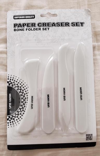 A set of 5" & 6" sized bone folders/paper creasers of different shapes stored in black and white branded packaging. On the left side of the packaging is a black circle with blobs surrounding its shape and the words "Morn Sun" printed within it. Bottom right corner underneath one of the bone folders is a mini QR code.