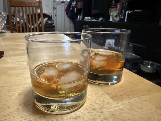 Two cocktail glasses with about an inch of an amber drink and ice cubes.