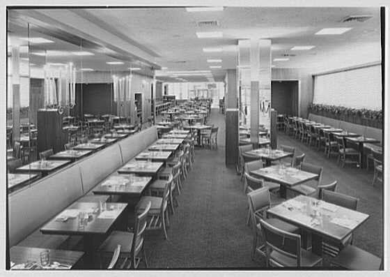 The image is a black and white photograph depicting the interior of what appears to be an Esso Building in Rockefeller Center, New York City. It shows a spacious dining area with multiple rows of square tables set up for service. Each table has placemats, glasses, silverware, and napkins neatly arranged on top.

The room is well-lit by large windows along the wall opposite where the photo was taken, allowing ample natural light to flood in. Reflective surfaces can be seen that suggest a mirror or glass partition running alongside one side of the dining area, reflecting part of the space and adding depth.

Chairs are placed around each table with no visible occupants at the moment the photograph was captured. The flooring is not clearly distinguishable but seems consistent throughout the room. There's an absence of people in the image which gives a sense of quietness or perhaps it’s taken during off-peak hours. Overall, this appears to be part of a general view within Schrafft's main dining area from June 9,1948 as suggested by the caption.

The quality and details suggest that this photograph is one among those collected in "Gottscho-Schleisner, Inc." collection which indicates it could have been used for commercial or architectural documentation purposes. The identifier provided points to a specific source of historical records related to New York City buildings during m [...]