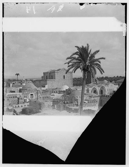 The image depicts a historical black and white photograph of an ancient town with stone buildings, indicative of Middle Eastern architecture. Prominent in the foreground is a tall palm tree standing against a partly cloudy sky. In the background lies what appears to be an older church or monastery made from large stones, featuring arched doorways and windows that speak to its historical significance. The setting suggests it may have been taken around 1900-1920 in Palestine, possibly Lydda (today's Lod) due to mention of the Church of St. George as part of "Jaffa to Jerusalem" context provided by reference information found online.