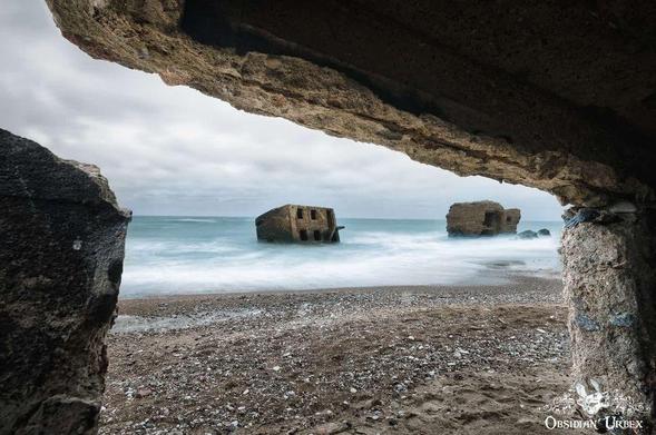 View from inside a concrete bunker looking out onto a beach with partially submerged, ruined bunkers in the sea under a cloudy sky.