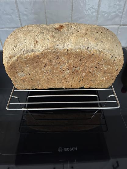 A loaf of home baked multigrain bread is sitting on a silver cooling rack, sitting on the black Bosch cooktop.  Some white splash back tiling is behind the loaf.