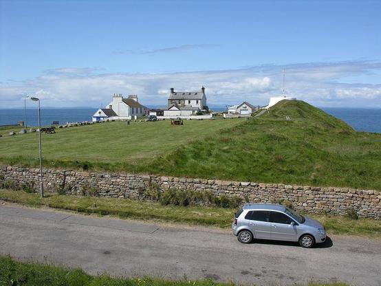 The remains of Burghead promontory fort at Burghead in Moray. The image shows a grassy area beyond a road and stone wall that cut across the bottom of the frame. There are buildings at the far end of the grassy area and a linear mound running along its right side, in line with the photographer. The scene is in sunlight.