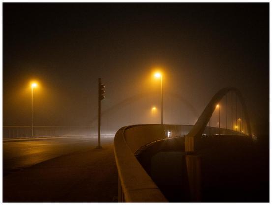 Blick auf eine Brücke bei Nacht, die Nebel eingehüllt und von Straßenlaternen beleuchtet ist.