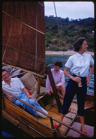 The image shows a group of people aboard a boat, with one woman standing and pouring herself a drink while smiling. The setting appears to be in Hong Kong during the summer months as indicated by the date on Extra 1959 reference. There are three individuals visible; two seated at the front end, where there's a cushioned area for comfort or relaxation.
The boat is equipped with sails made of what looks like brown fabric tied up and not extended fully. The water appears calm in this photograph captured by Toni Frissell for Sports Illustrated magazine.