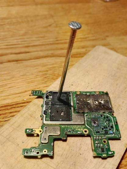 A board with a qualcomm mobile chipset on a small sheet of plywood with visibly damaged chips. The main SoC is pierced by a big nail,