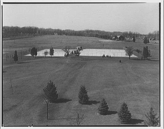 The image is a black and white photograph depicting an expansive outdoor scene, likely taken during the early 20th century. In this wide-angle view, there's a large, fenced-in ice rink in the center that appears to be frozen over with several people around it engaged in various activities or socializing near its perimeter.

The foreground features several trees and what seems like young saplings planted at intervals across open fields. The middle ground is mostly flat expanse of grass leading up to the fence surrounding the rink, which acts as a natural barrier separating this area from other buildings visible further away.

In the background, beyond the ice skating field, there are some farm-like structures or barns with large doors and what seems like a collection of storage containers. The scene is set in an open countryside environment characterized by vast fields that seem to extend towards distant trees marking the horizon line. This setting indicates rural surroundings rather than urban landscapes.

There's no visible sign of modern amenities, suggesting this might be an old-time snapshot capturing daily life or leisure activities from a past era at Georgetown Preparatory School based on provided context. The image evokes a sense of tranquility and simplicity common in historical scenes before the advent of extensive technological advancements and infrastructural d [...]