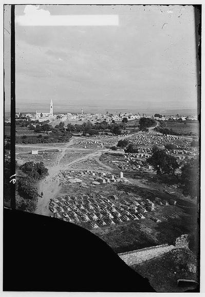 The image is a black and white photograph showing an aerial view of what appears to be a town with various structures including buildings, a church or tall building resembling a minaret. Below the town lies fields and clusters of graves in large cemeteries organized in rows. The perspective suggests that this photo may have been taken from above using a camera on a tower or another elevated structure. It's a historical snapshot likely documenting urban development during an early 20th-century time period, as indicated by the style of buildings, infrastructure, and absence of modern vehicles.