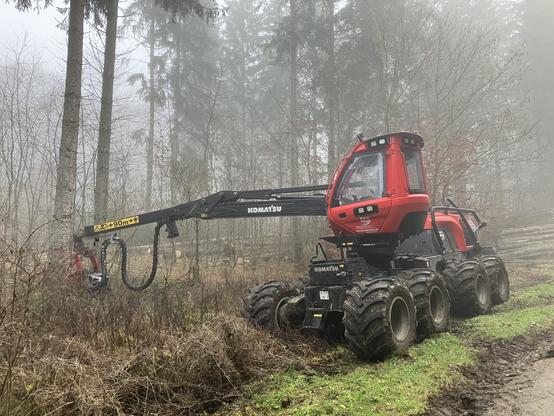 Eine rote Forstmaschine steht im Wald sie hat einen langen schwarzen Ausleger. Auf dem steht ein gelbes Schild mit einem Piktogramm, dass man 90 m Abstand halten soll.