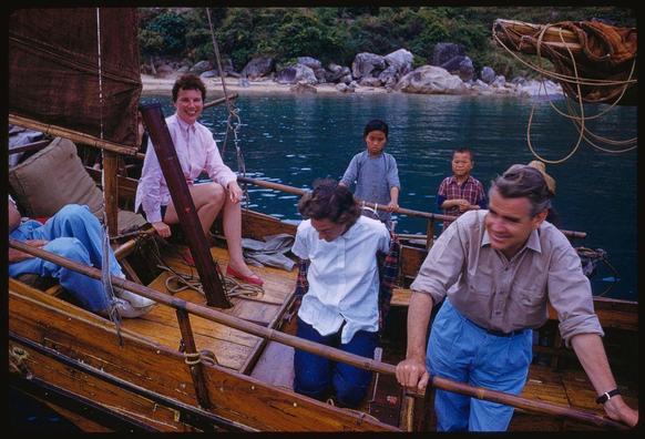 Five people are on a wooden boat in calm water. Two women, one sitting and smiling holding what appears to be a folded umbrella or similar item, the other standing near her with legs crossed wearing blue pants and red shoes; two young boys stand beside them while another woman sits down turned away from view but still looking at something outside of camera range; all five appear cheerful. The boat has ropes attached and there are rocks in the background as well as trees on a shoreline that appears to be part of Hong Kong, based on caption information provided by LOENER.