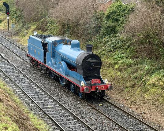 A diagonal side view from a high point of view of a powder blue steam locomotive with red running boards, two small leading wheels, two big driving wheels and a tender well filled with coal.
