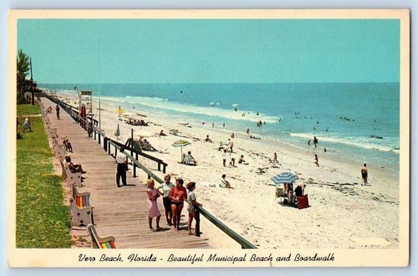 This vintage postcard shows the municipal beach and boardwalk at Vero Beach, Florida, likely mid-20th century. The wooden boardwalk runs parallel to the shoreline, lined with painted benches and a low railing. People stroll along the walkway while others relax on the sand under striped umbrellas or sunbathe closer to the water. Swimmers are visible in the gentle surf, and the wide turquoise horizon emphasizes the Atlantic Coast setting. The scene captures a classic Florida beachfront—clean sand, bright sun, and a leisurely atmosphere typical of postwar seaside tourism.