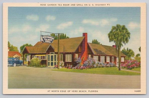 This vintage linen-style postcard shows the Rose Garden Tea Room & Grill on U.S. Highway 1 at the north edge of Vero Beach, Florida. The single-story building features a steeply pitched roof, wood siding, a prominent brick chimney, and a glazed entry vestibule. Flower beds—likely roses—border the façade and a palm tree marks the right-hand lawn, reinforcing the Florida setting. A painted hanging sign above the entrance reads “Rose Garden Tea Room & Grill.” A period automobile is partially visible at left, helping date the image to the mid-20th century. The composition emphasizes roadside hospitality and gardened landscaping typical of small coastal eateries promoted to motorists on historic U.S. highways.
