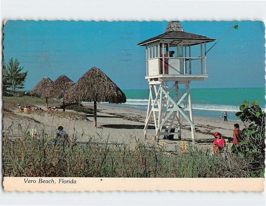 This color postcard shows a coastal scene at Vero Beach, Florida, likely from the late 1960s or early 1970s. A tall white wooden lifeguard tower stands prominently in the foreground, featuring cross-braced legs, a small enclosed lookout cabin, and a shingled hip roof. A green flag is raised on a short pole attached to the tower, indicating favorable swimming conditions. To the left, several palapa-style thatched beach shelters line the shoreline, with visitors relaxing beneath them. Farther down the beach, people walk and play near the surf. The Atlantic Ocean appears calm with low waves, set against a bright turquoise horizon and a cloudless sky. Coastal vegetation, including sea oats and low shrubs, frames the lower portion of the image. The caption reads “Vero Beach, Florida.”