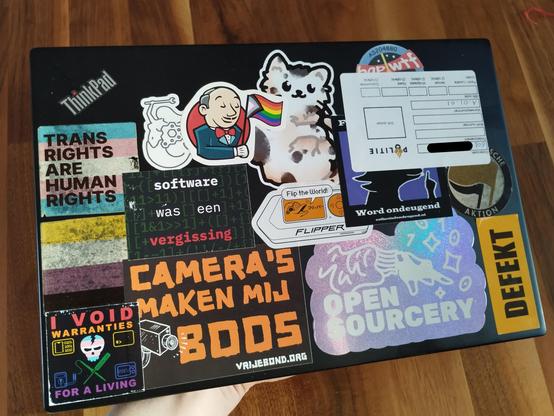A ThinkPad cover, full of various stickers. On top of a "Fuck de status quo, word ondeugend", Antifaschistische Aktion, and bgp.wtf, there is an upside down sticker with the logo of Politie, and a case signature starting "EOB-" handwritten with a pen