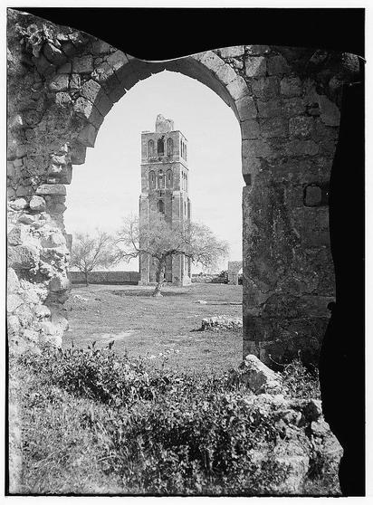 The image depicts a historical scene captured through an arched stone doorway, providing a view of the Tower of Forty Martyrs in Ramleh. The tower stands prominently within what appears to be ruins or an old church structure, with visible Gothic architectural elements such as pointed arches and tall narrow windows. Surrounding the site is grassy terrain with scattered bushes, hinting at natural overgrowth typical of abandoned historical sites.

The photograph's monochromatic tone suggests it may have been taken in the early 20th century or earlier, indicative of its age and possibly a documentary nature capturing the ruins during that period. The framing through an archway adds to the sense of viewing history from another time, emphasizing both the passage of time since these structures were fully intact as well as their historical significance.

Additional context is provided by reference information indicating this photograph was part of "Jaffa to Jerusalem," likely a collection or publication about various locations in Israel and Palestine. The mention of American Colony (Jerusalem) Photo Department suggests that it could have been taken within the premises of an institution with connections to photography, possibly associated with Christian missionary activities during late 19th-early 20th centuries.

The reference number "691e" along with a unique identifier "[approx [...]