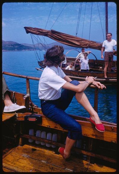 The image depicts a woman with shoulder-length brown hair wearing red shoes and dark blue pants, sitting on the deck of a wooden boat. She is relaxedly resting her legs against one another while looking out at something beyond. Another individual stands in the background holding what appears to be an object that could potentially be identified as smoke. The sea behind them indicates they are not far from shore with land visible in the distance, and there's also a glimpse of more boats or ships on the water nearby.