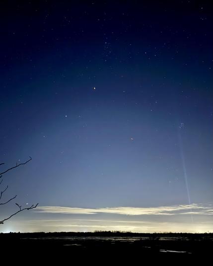 A wide view of a starry sky over a rural scene with puddles of water reflecting the band of light pollution at the horizon. A beam of light points up from the horizon into a cluster of stars.
