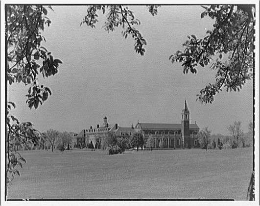 The image is a black and white photograph depicting an expansive, historical-looking building with multiple wings and structures. In the foreground are branches from trees framing the top left corner of the picture. The main structure appears to be made up of brick or stone blocks and features prominent architectural elements such as steeply pitched roofs, arched windows, ornate trimmings along edges, a clock tower in the center right with spires reaching upwards, and what seems like chimneys protruding from the roofline.

The building is situated on an open field that extends toward the horizon. There are no visible people or vehicles around; it's devoid of any significant activity which might suggest this could be either early morning before anyone arrives for classes or perhaps a special event where visitors have been cleared out to provide better visibility in the photograph. The overcast sky gives off a muted, serene atmosphere that complements the stately and somewhat grandiose nature of Georgetown Preparatory School as it was likely captured during its formative years.

This image is tagged with "Horydczak" which indicates the photographer's name or their associated pseudonym and dates from around 1890 to 1971, suggesting that this photograph might be over a century old. It has been digitally preserved by Loener NL under the reference '691e/691ec93c450e57a2674d2a87 [...]