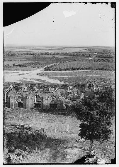 The image is a black and white photograph depicting an expansive rural landscape with remnants of ancient architecture. In the foreground, there's a dilapidated stone structure featuring arches, indicative of Roman or Byzantine construction. The ruins are overgrown with vegetation, suggesting they have been abandoned for some time.
Beyond this ruinous edifice lies rolling farmland divided into neat plots by winding paths and irrigation ditches, hinting at agricultural activity. A river cuts through the center of the scene from left to right, its banks lined with sparse trees or shrubs. The landscape extends towards a horizon marked by an open plain under what appears to be overcast skies.
In one corner of the photograph stands a solitary tree with twisted branches and dense foliage on top, which adds contrast against the flat expanse of fields in the distance. There are no visible people in this image; instead, it focuses on the juxtaposition between nature's resilience through ancient ruins amid cultivated land under an unassuming sky.
This black-and-white photo may have been taken during a historical period when such imagery was common for documenting landscapes and architecture with less emphasis on color photography which became popular later.