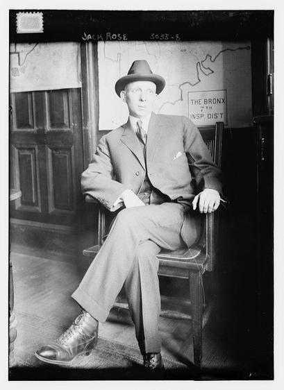 The image is a black-and-white photograph of a man seated in a wooden chair. The man is dressed formally in a suit with a tie and a hat, suggesting a professional or formal setting. He is wearing dress shoes and has a handkerchief in his breast pocket. His posture is relaxed, with one hand resting on the chair's armrest. The background features a wooden door and a wall with a map and a sign that reads "THE BRONX 7TH INS.P. DIST." The photograph appears to be historical, given the style of the clothing and the quality of the image. The man is identified as "Jack Rose" based on the text at the top of the image. The overall setting suggests an office or administrative environment.
