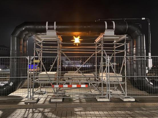 Ein Dehnungsbogen einer Fernwärmeleitung hinter einem Bauzaun. Ein leuchtender Herrnhuter Stern hängt unter dem Bogen.