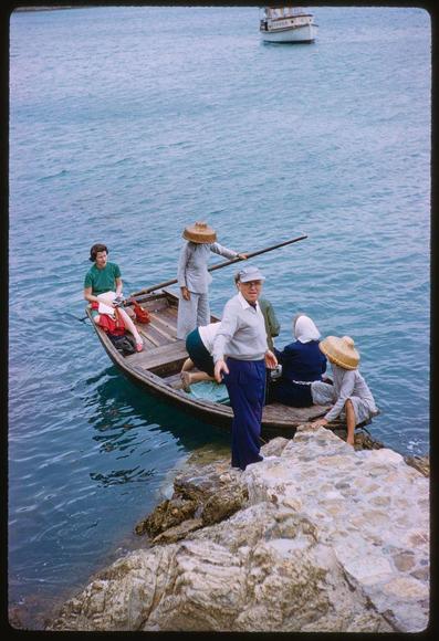 A color photograph showing a group of people on a small boat in the water, likely engaged in recreational boating. The individuals appear to be tourists or sightseers, as suggested by their casual attire and poses for the photo.

In the foreground is an older man wearing blue pants, white shirt, cap, and straw hat standing with his right arm extended towards another person on a rock formation beside him; he seems to be guiding or interacting in some manner. The next group of people are seated on the boat: two individuals appear to be tourists, one dressed casually and sitting while the other is partially obscured but appears to be handling equipment.

Onboard there's also an individual wearing red shoes, a green shirt, white cap, kneeling with their legs spread apart as if posing for the picture. Behind them stands another person in dark pants and blue jacket looking towards something outside of frame or perhaps interacting with someone not pictured here.
In contrast, on land is a larger rock formation that appears to be part of a beachside setting where other individuals are seated casually.

The water surrounding the boat shows gentle waves indicating calm seas. Above them floats another vessel which could indicate it's near an area frequented by boats or perhaps heading towards one as they might have embarked from there earlier.
Overall, this scene captures leisure and [...]