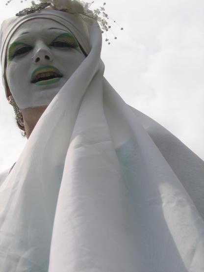 A person with white face paint and green and yellow eye shadow and lipstick wearing a white head scarf topped with a broach and small flowers. The shot is from below, looking up at their face at the top left of the shot, and the scarf swoops down from the side of their head to fan out dramatically towards the bottom of the shot. The sky behind them is white with clouds, so the entire shot is almost monochrome, save for their eyes and lips.