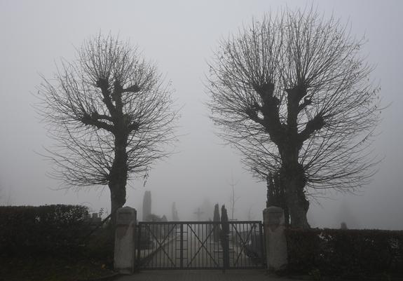 Zwei blattlose Bäume, die ein geschlossenes Tor flankieren, dahinter liegt alles im Nebel, zu erkennen ist ein Weg, ein Kreuz und einzelne Grabmäler, was auf einen Friedhof  hindeutet.